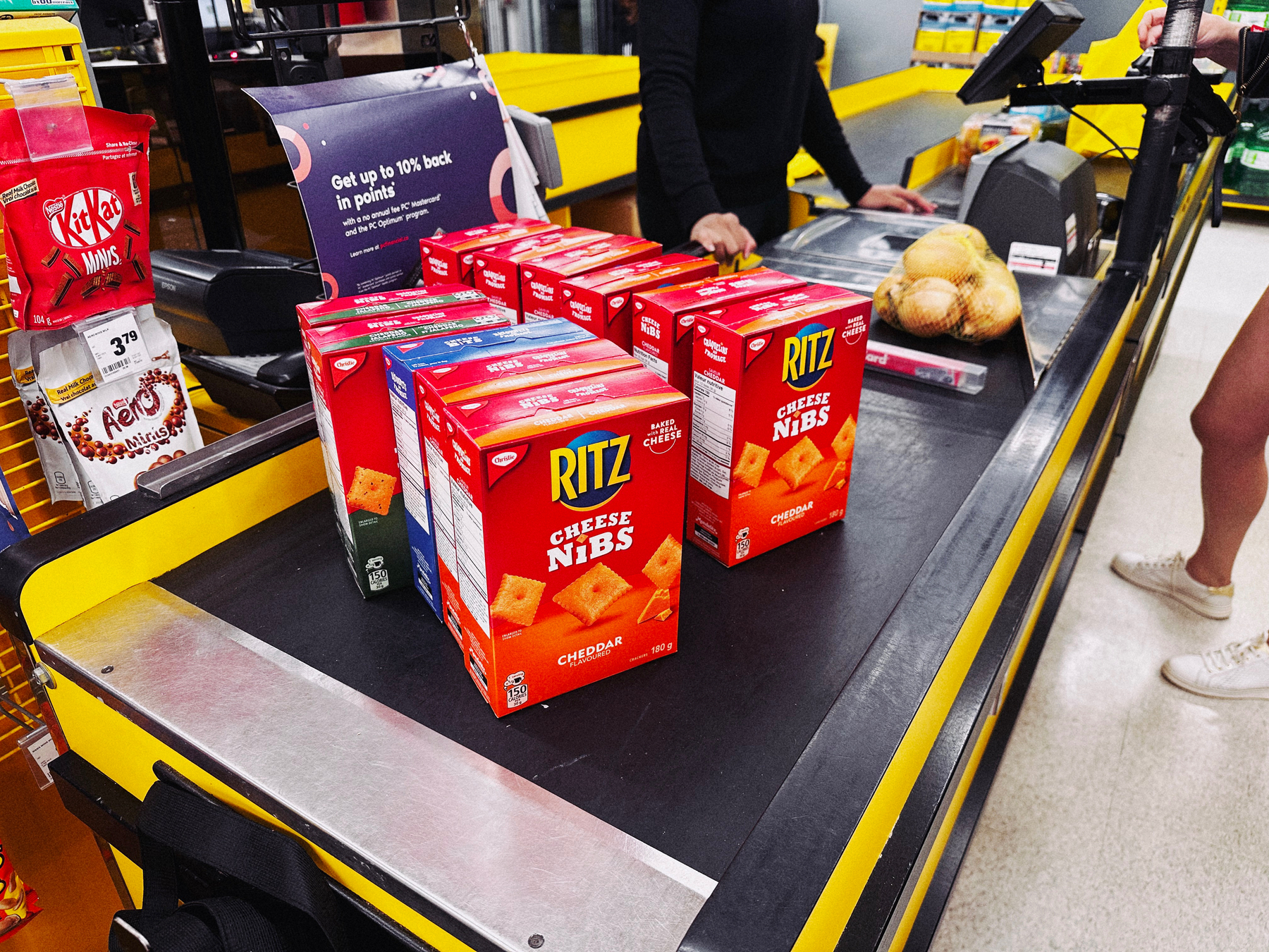 11 boxes of Ritz Cheese Nibs on a grocery store checkout conveyor. 